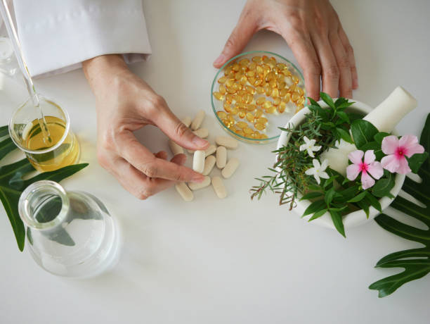 Top-down view of hands handling yellow capsules from a small bowl beside a glass of water and a flowering plant on the table